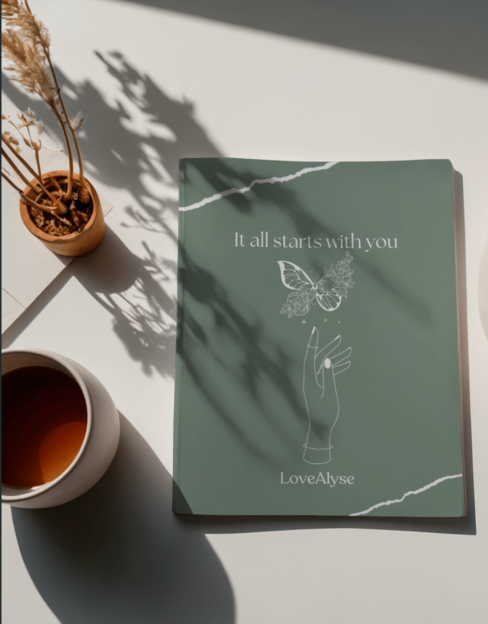 Journal titled 'It all starts with you' by LoveAlyse on a white surface with a cup and plant.